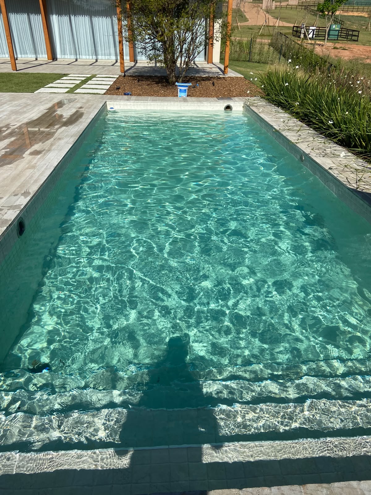 piscina retangular de água verde-esmeralda com deck de pedra clara, pergolado de madeira e jardim ao fundo  — texto:  