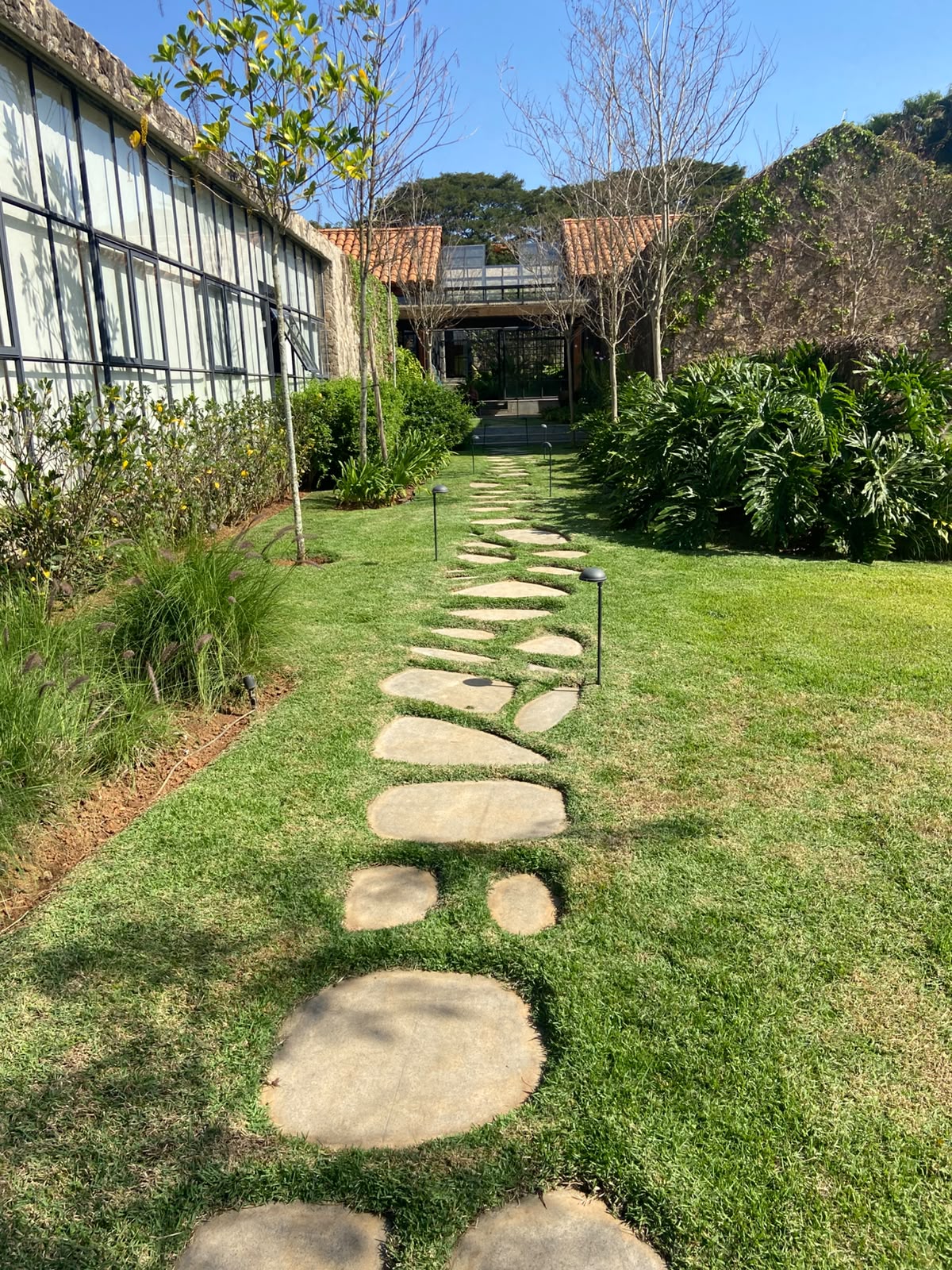 caminho de pedras irregulares sobre gramado em jardim paisagístico, com casas de telhado de telha e canteiros 