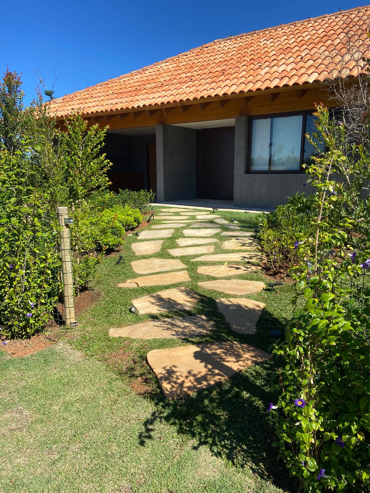 caminho de pedras irregulares sobre grama levando à entrada de uma casa com telhado de telha colonial e jardim